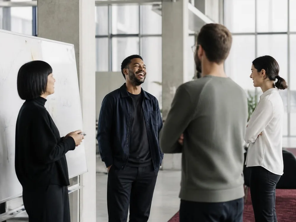 Four colleagues hold a relaxed stand-up by a whiteboard in a light-filled office, one of them laughing.